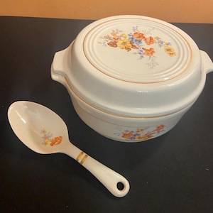 May include: White ceramic casserole dish with lid and serving spoon. The dish and spoon are decorated with a floral pattern in orange, yellow, and blue. Gold trim accents the edges of the dish and spoon handle.