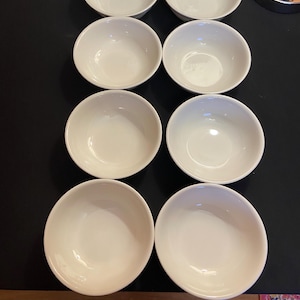 May include: Eight off-white ceramic bowls arranged in two rows of four. The bowls are round with a simple, classic design. The bowls are likely for serving soup, cereal, or other dishes. The bowls are on a dark surface.