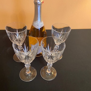 May include: A champagne bottle with a silver foil top and a label, surrounded by four crystal wine glasses. The glasses have a detailed, cut-glass design.