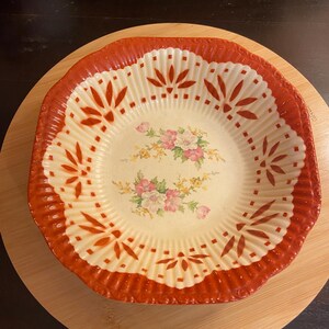 May include: A vintage, cream-colored ceramic bowl with a scalloped, rust-colored rim. The bowl features a floral design in the center and decorative leaf patterns around the rim. The bowl is sitting on a wooden surface.