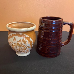 May include: Two ceramic vessels. One is a small, round, orange pot with a white floral design. The other is a tall, dark brown mug with a handle and a textured surface. Both are on a black surface.