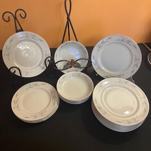 May include: A collection of white ceramic dinnerware with a silver leaf design and gold trim. The set includes plates and bowls, displayed on decorative black metal stands. The plates are arranged on a black surface.