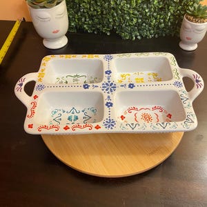 May include: A white ceramic serving tray with four rectangular compartments and handles. The tray is decorated with colorful floral and geometric patterns in red, blue, yellow, and green. It sits on a wooden lazy susan.