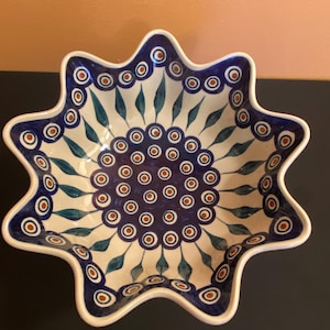 May include: A star-shaped ceramic bowl with a white base and a blue and brown floral pattern. The bowl has a scalloped edge and is likely used for serving food or as a decorative piece. The design includes blue teardrop shapes and brown circles.