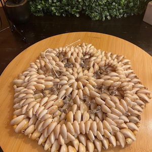 May include: A hexagonal trivet made of numerous small, cream-colored seashells. The shells are arranged closely together, creating a textured surface. The trivet is on a round, light-brown wooden surface.