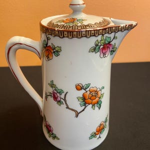 May include: White ceramic creamer with a lid, adorned with orange, pink, and green floral patterns. The creamer has a handle and a spout. The lid is topped with a small knob. The rim of the creamer and lid are decorated with a brown border.