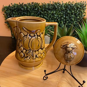 May include: A tan ceramic pitcher with a raised relief design of grapes and leaves on the body. The matching circular lid features a sculpted fruit finial and rests on a black metal stand. The pitcher has a handle and a pouring spout.