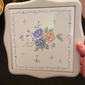 May include: A square, off-white trivet with a floral design. The center tile features a bouquet of blue and peach roses with green leaves. A light blue border with small floral accents surrounds the central design. The trivet has a light gray wooden frame.