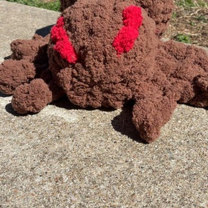 Könnte beinhalten: Ein großes, handgefertigtes, braunes Häkel-Spinne-Plüschtier mit roten Akzenten. Die Spinne hat einen runden Körper und mehrere Beine, alle aus einem weichen, strukturierten Garn. Das Plüschtier liegt auf einer Betonoberfläche.