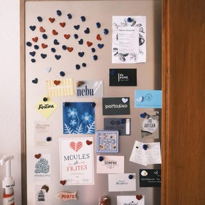 Könnte beinhalten: Eine rechteckige magnetische Pinnwand mit Holzrahmen, bestückt mit zahlreichen kleinen herzförmigen und runden Magneten in Rot und Blau. Verschiedene Karten, Menüs und Notizen sind befestigt, darunter der Text Moules et Frites.