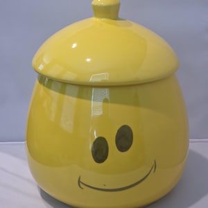 May include: A bright yellow ceramic jar with a matching lid featuring a knob handle. The jar has a rounded, bulbous shape and displays a dark gray graphic of two oval eyes and a curved smiling mouth on the front surface of the container.