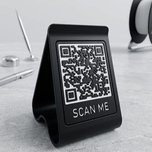 May include: A black QR code display stand with the text "SCAN ME" in white. The stand is triangular and sits on a grey surface. The QR code is black and white.