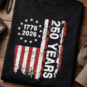 Pu&ograve; includere: T-shirt nera con un design invecchiato della bandiera americana. Il design include il testo "1776 2026" e "250 YEARS". La bandiera presenta strisce rosse e bianche e stelle bianche, celebrando un anniversario significativo.