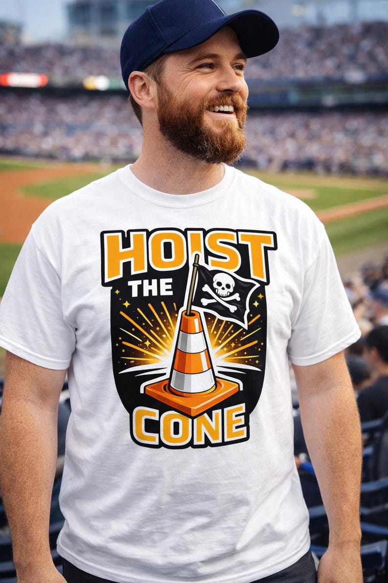 Puede incluir: Camiseta blanca con el texto "HOIST THE CONE" en naranja y negro. El dise&ntilde;o presenta un cono de tr&aacute;fico con una bandera pirata. La persona de la imagen lleva una gorra de b&eacute;isbol azul marino.