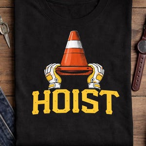 Puede incluir: Camiseta negra con un gr&aacute;fico de un cono de tr&aacute;fico naranja y blanco sostenido por manos enguantadas, con la palabra "HOIST" en letras amarillas. El dise&ntilde;o est&aacute; centrado en la camiseta.