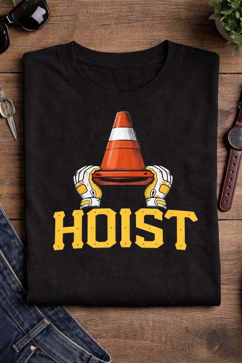Puede incluir: Camiseta negra con un gr&aacute;fico de un cono de tr&aacute;fico naranja y blanco sostenido por manos enguantadas, con la palabra "HOIST" en letras amarillas. El dise&ntilde;o est&aacute; centrado en la camiseta.