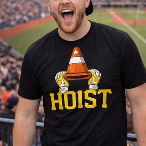 Puede incluir: Camiseta negra con un gr&aacute;fico de un cono de tr&aacute;fico naranja y blanco sostenido por manos enguantadas, con la palabra "HOIST" en amarillo. Un hombre con una gorra de b&eacute;isbol azul sonr&iacute;e en el fondo.