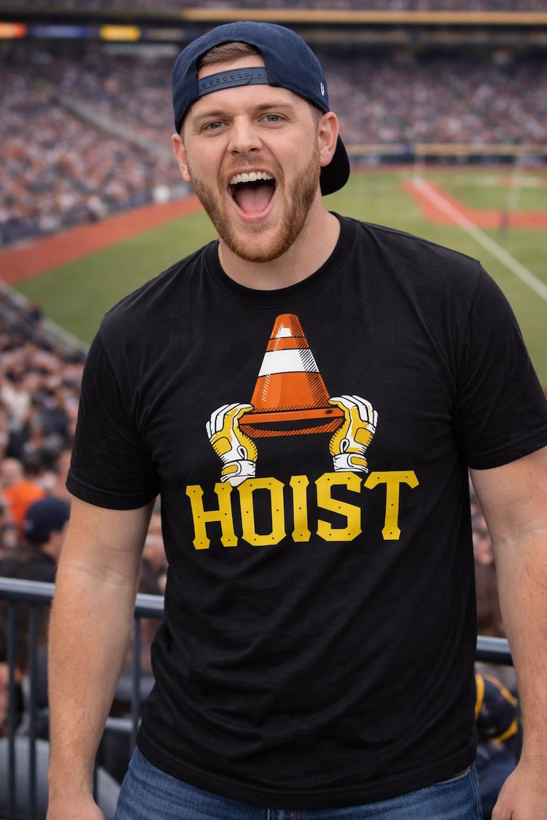 Puede incluir: Camiseta negra con un gr&aacute;fico de un cono de tr&aacute;fico naranja y blanco sostenido por manos enguantadas, con la palabra "HOIST" en amarillo. Un hombre con una gorra de b&eacute;isbol azul sonr&iacute;e en el fondo.