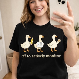 May include: Black t-shirt featuring a design of three ducks, each with an item: a pencil, a shirt with text, and a coffee cup. The text "off to actively monitor" is printed below the ducks.