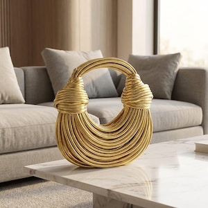 Pode incluir: Uma bolsa dourada com um design único. A bolsa apresenta uma alça em laço e um corpo composto por inúmeros fios paralelos e curvos. A bolsa está sobre uma mesa de mármore em uma sala bem iluminada.