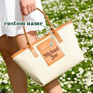 May include: Cream-coloured tote bag with brown leather accents and handles. The bag features a brown leather patch with the name "Michael Joe" embossed on it. The bag is held by a person in a white dress.
