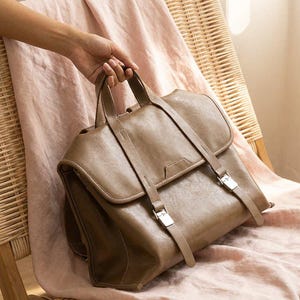 Puede incluir: Una mochila de cuero marrón con cierre de solapa y correas ajustables. La bolsa tiene hebillas plateadas y un asa superior. Descansa sobre una tela rosa sobre una silla de mimbre.