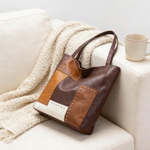 May include: A brown and tan tote bag with a patchwork design and studded accents. The bag has two handles and is resting on a white couch next to a beige mug and a cream-colored blanket.