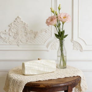 May include: A cream-colored clutch with a gold clasp rests on a round wooden table covered with a cream-colored lace doily. A clear glass vase holds pink flowers with green stems. The background features white wall molding.