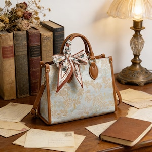 May include: A light blue and beige tote bag with a landscape pattern, brown handles, and a decorative scarf. The bag has a detachable shoulder strap and is sitting on a wooden surface.