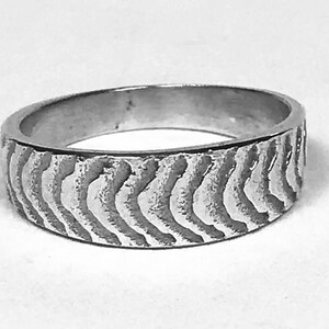 Könnte beinhalten: Ein silberner Ring mit einem strukturierten, wellenartigen Muster um das Band. Der Ring hat einen einfachen Bandstil mit leicht abgerundeter Innen- und Außenseite. Der Ring wird vor einem weißen Hintergrund gezeigt.