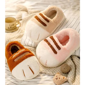 May include: Three pairs of plush, cat paw-shaped slippers in the image. One pair is brown and orange, another is pink, and the third is beige. Each slipper has a white paw print design on the toe and brown stripes across the top. The slippers are on a striped blanket.