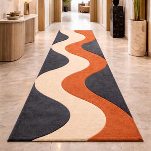 May include: A long, rectangular rug with a wavy design in shades of dark gray, cream, and orange. The rug is placed in a hallway with a marble floor. The design is a modern, abstract pattern.
