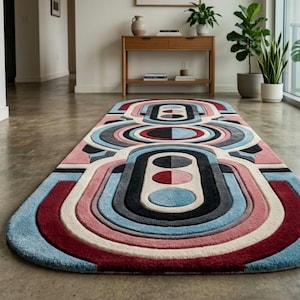 May include: A rectangular rug with a rounded end features a geometric design in shades of red, pink, blue, gray, and black. The rug is placed on a concrete floor, with a view of a hallway and a window with plants.