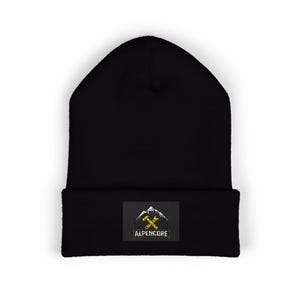 Op de afbeelding: Zwarte beanie met een rechthoekige patch. De patch toont een berglandsilhouet, gekruiste pikhouwelen en het woord "ALPENCORE" in geel, met een verweerde lettertype. De beanie is gemaakt van zacht gebreid materiaal.