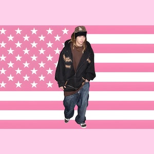 Könnte beinhalten: Eine Person trägt eine schwarze Kapuzenjacke mit braunen Aufnähern, ein braunes Hemd und eine weite blaue Jeans. Sie trägt eine gemusterte Baseballkappe und steht vor einem rosa-weiß gestreiften Hintergrund mit weißen Sternen.