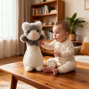 May include: A plush stuffed animal ferret toy with gray, white, and brown fur, a pink nose, and black eyes. The toy is sitting on a wooden table next to a baby wearing a cream-colored onesie.
