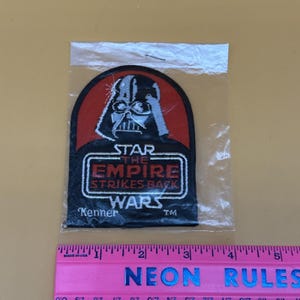 Op de afbeelding: Een Star Wars-patch met Darth Vader in een zwart en rood ontwerp. De patch leest "STAR THE EMPIRE STRIKES BACK WARS" met het Kenner-logo. De patch zit in een doorzichtige plastic zak, met een roze liniaal eronder.