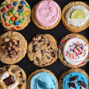 May include: Nine assorted cookies on a black tray. The cookies are decorated with colorful candies, pink frosting, a lime slice, chopped peanut butter cups, chocolate chips, sprinkles, a marshmallow and chocolate bar, a blue gummy shark, and Oreo cookies.