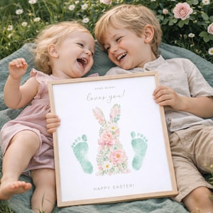 Könnte beinhalten: Ein gerahmter Druck mit einem hellen Holzrahmen, der von zwei Kindern gehalten wird. Der Druck zeigt ein Kaninchen aus Blumen und Fußabdrücken mit dem Text "SOME BUNNY loves you!" und "HAPPY EASTER!" in schwarzer Schrift. Die Kinder lächeln.