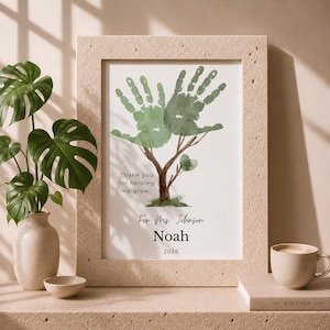 Könnte beinhalten: Ein gerahmter Druck mit einer Baumillustration, bei der grüne Handabdrücke als Blätter dienen. Der Text lautet: Thank you for helping me grow. For Ms. Johnson, Noah, 2026. Der Rahmen hat eine strukturierte, hellbeige Oberfläche.