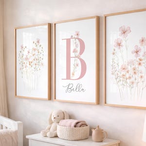 May include: Three framed wall art prints in light wood frames. The left and right prints feature watercolour-style pink and white wildflowers. The centre print displays a large pink letter B with the name Bella written below in black script.