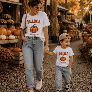 Puede incluir: Dos camisetas blancas, una con el texto MAMA y la otra con MINI, ambas con un gráfico de una calabaza naranja y la palabra Pumpkin impresa debajo. Las camisetas se combinan con pantalones vaqueros de mezclilla azul claro y zapatillas.