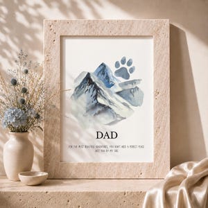 Könnte beinhalten: Ein gerahmter Druck mit einer Aquarellillustration von blauen Bergen und einem grauen Pfotenabdruck. Der Text lautet: DAD. FOR THE MOST BEAUTIFUL ADVENTURES, YOU DON'T NEED A PERFECT PLACE. JUST YOU BY MY SIDE. Der Rahmen hat eine strukturierte, beige Oberfläche.