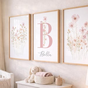 Könnte beinhalten: Drei gerahmte Kunstdrucke mit floralen Motiven und einer hellen Holzoberfläche. Die Drucke zeigen Aquarellillustrationen von rosa Blumen und Grün. Der mittlere Druck hat einen großen rosa Buchstaben "B" mit dem Namen "Bella".