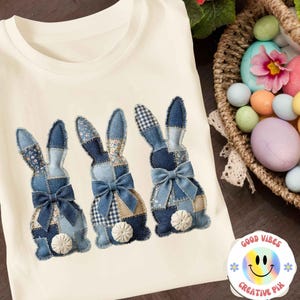 Puede incluir: Camiseta color crema con tres diseños de conejos de patchwork de mezclilla con lazos azules y colas de pompón blancas. Los conejos están hechos de varios patrones de mezclilla. Una cesta de huevos de Pascua está en el fondo.