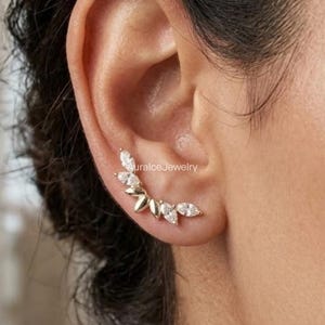 May include: A gold ear climber earring featuring a curved row of marquise-cut clear stones that follow the shape of the earlobe. The piece displays a metallic gold finish and includes the text AuralceJewelry printed in small letters near the center.