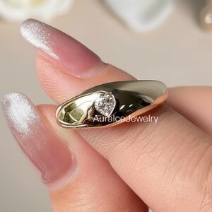 Puede incluir: Un anillo dorado con un diamante en forma de corazón. El anillo tiene un diseño liso y redondeado. El texto "Auralcojewelry" es visible en el anillo. El anillo está sostenido por una persona.