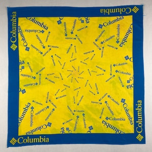 Könnte beinhalten: Ein quadratisches gelbes Bandana mit blauem Rand. Das Bandana ist mit dem Wort "Columbia" in Blau sowie dem Columbia-Logo bedruckt. Der Text und das Logo wiederholen sich im gelben Zentrum.