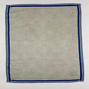 Peut inclure: Un mouchoir carré gris clair avec un motif quadrillé. Les bords sont bordés d'une bordure gris foncé, d'une bande bleue et d'une bande blanche. Les mots "PIERRE BALMAIN" sont imprimés en bas à droite.