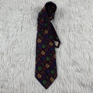 May include: A navy blue tie featuring a floral design in yellow, green, and blue. The tie is made of a smooth material and is displayed on a white, fluffy surface. The tie is rolled at the top, showing the pattern.